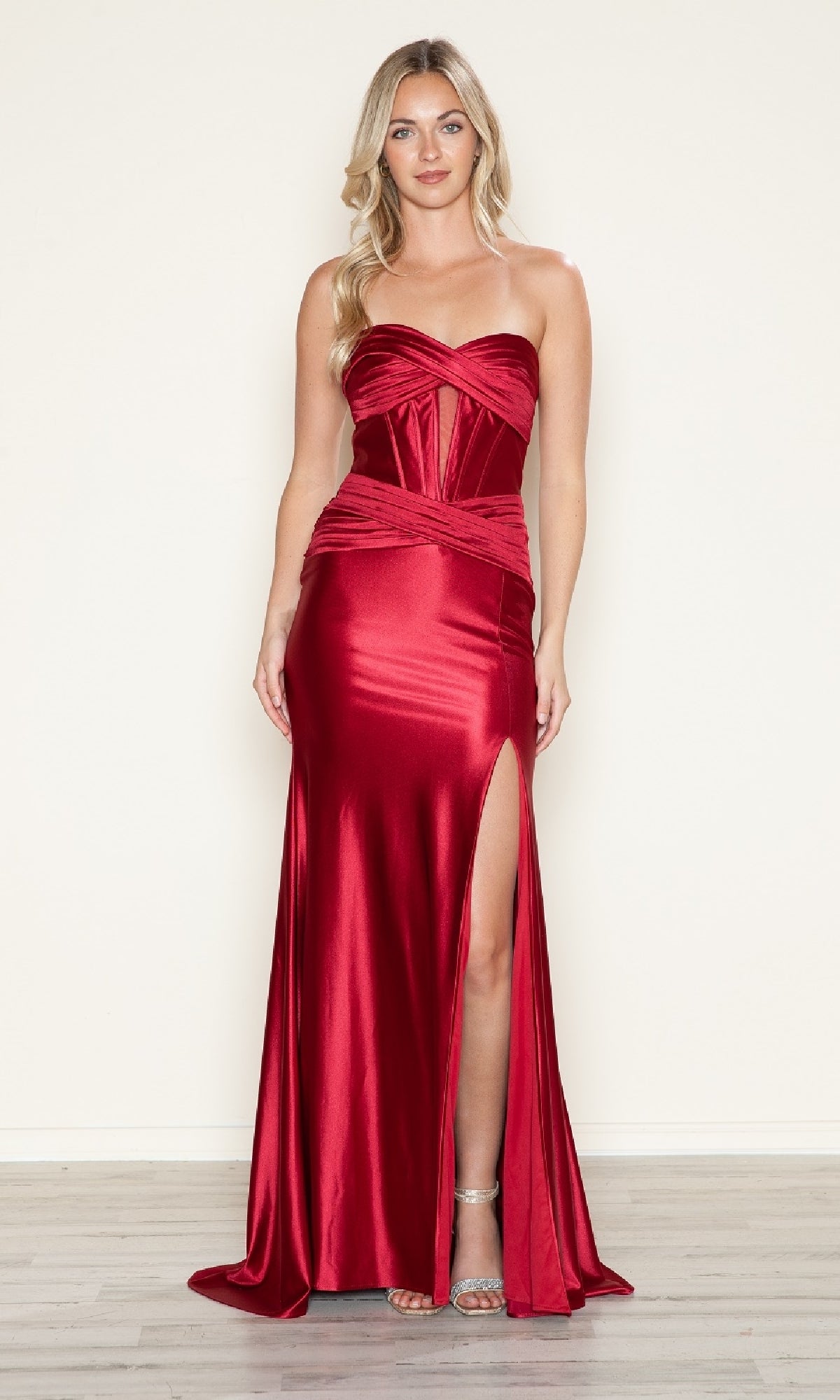 A woman in the Strapless Keyhole Long Prom Dress: Poly USA 9574, featuring a corset-style bodice and high side slit, stands on a light wood floor against a plain light background.